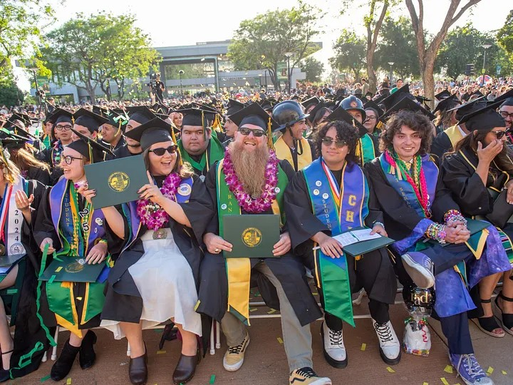 A group of happy graduates sit together outdoors in caps and gowns at the Craftonhills 2025 commencement, holding diplomas. They're adorned with leis and sashes, celebrating their achievement.